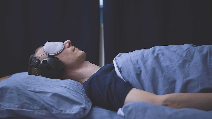 A man lying down on a bed in a dark room with an eyemask and headphones on