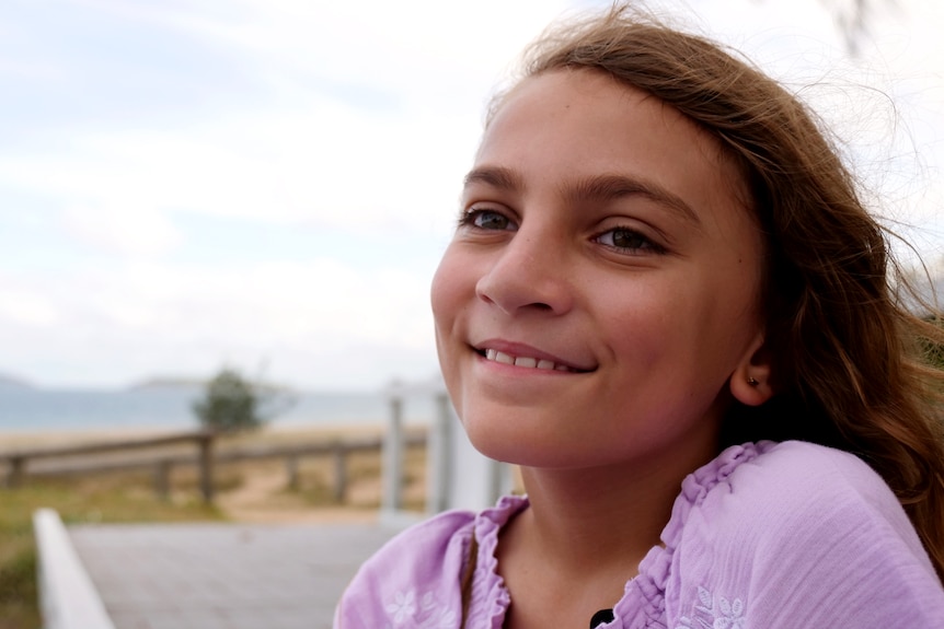 a young girl with long hair smiles at the camera, she is wearing a purple dress and sitting at the beach