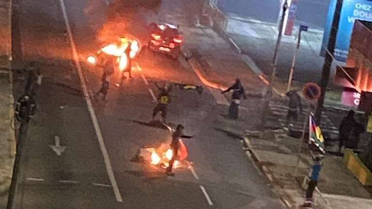 A nighttime photo of people rioting in the road. There's things on fire on the road and people standing around.