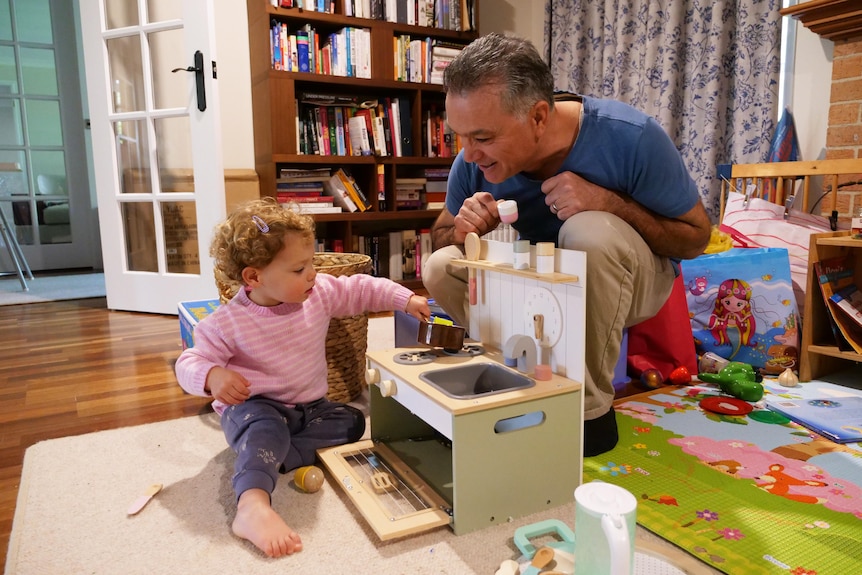 A man squatting watches a small girl play with a miniature toy kitchen