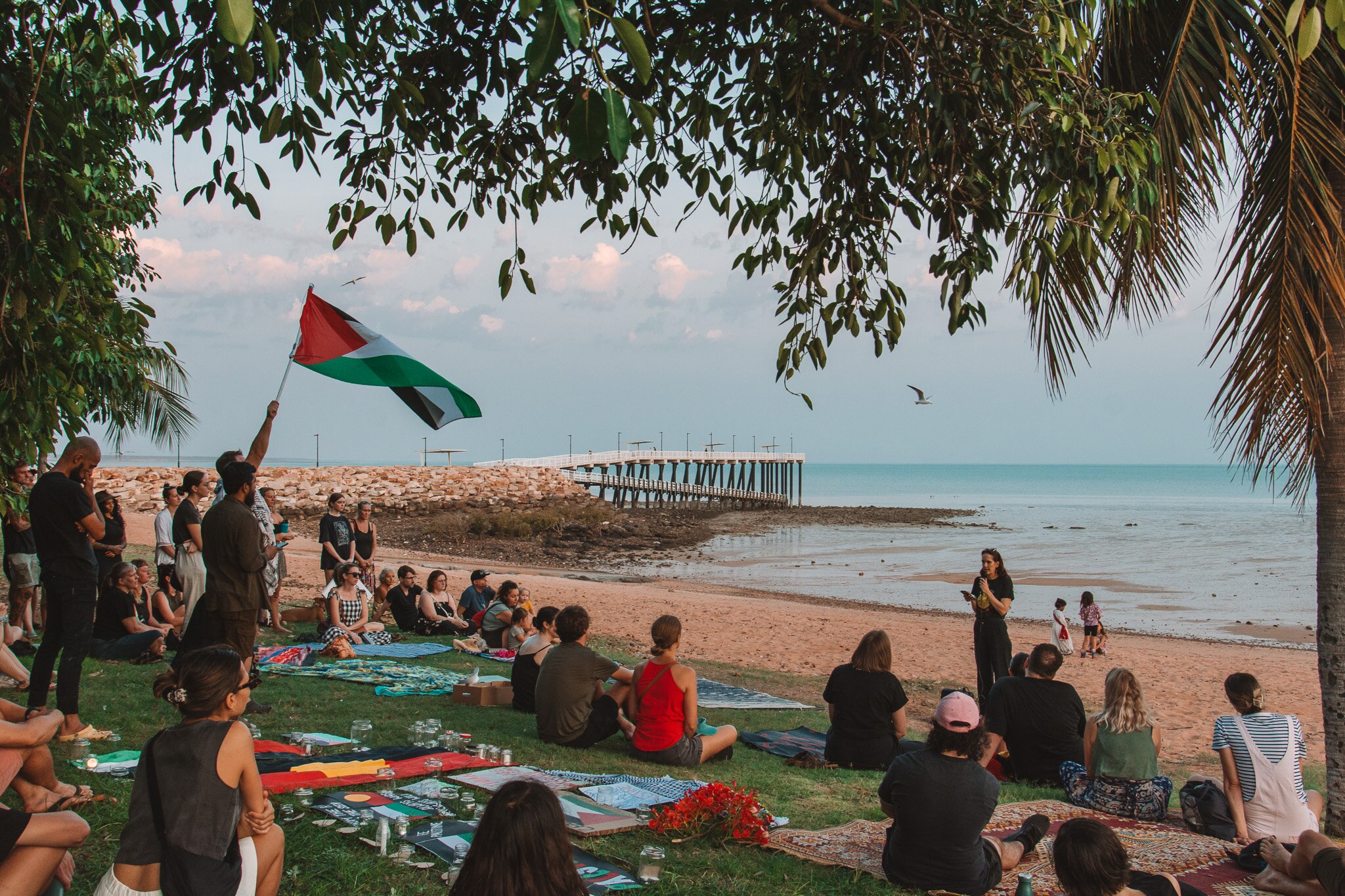 A group of people, one holding a Palestinian flag, sitting on grass in front of a woman standing on the beach with a microphone.