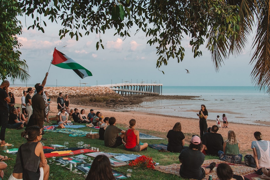 A group of people, one holding a Palestinian flag, sitting on grass in front of a woman standing on the beach with a microphone.
