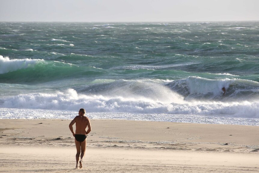 A man in speedos jogs towards the rough City Beach water as a storm approaches. Another body surfs.