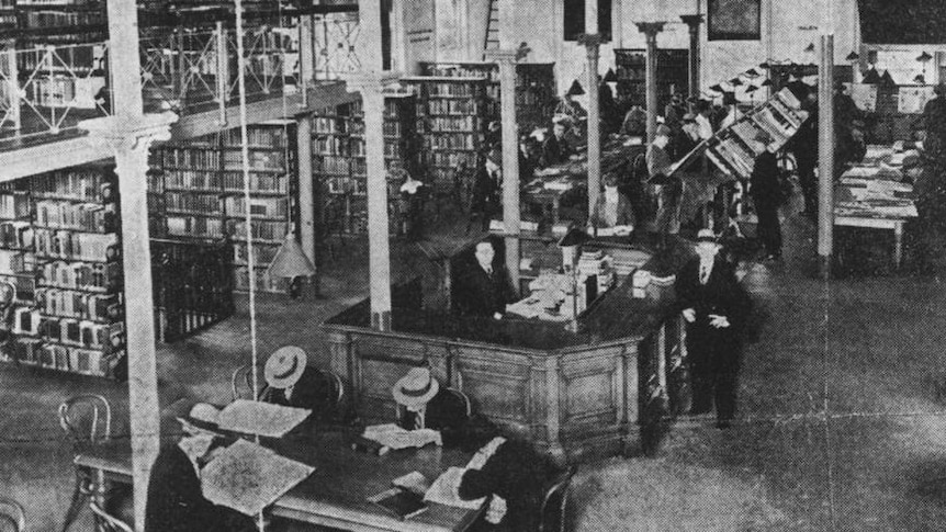 Black and white photo of a men reading papers in a room full of books.