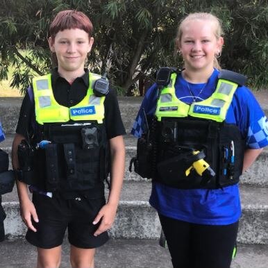 Three students stand smiling at camera wearing high vis police uniform vests
