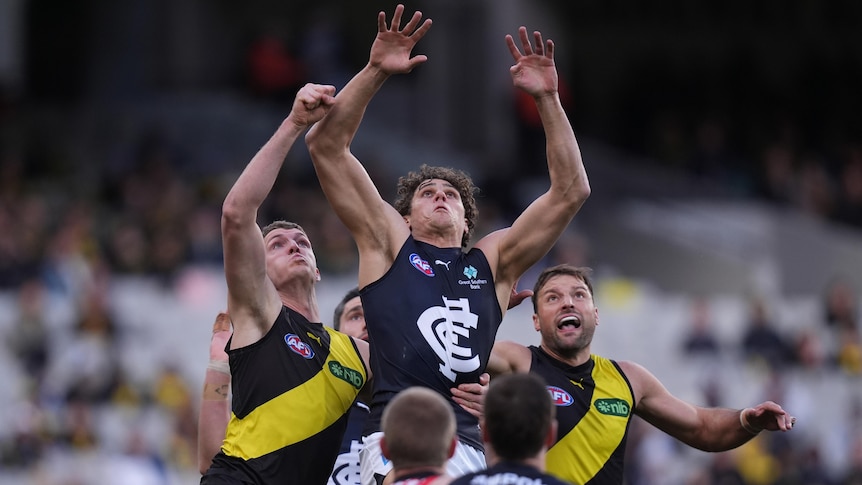 A Carlton AFL forward grimaces as he reaches up to try to mark the ball surrounded by Richmond defenders.