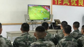 Rows of young men in camouglage and buzzcuts sit in a room, watching a football game on a large television.