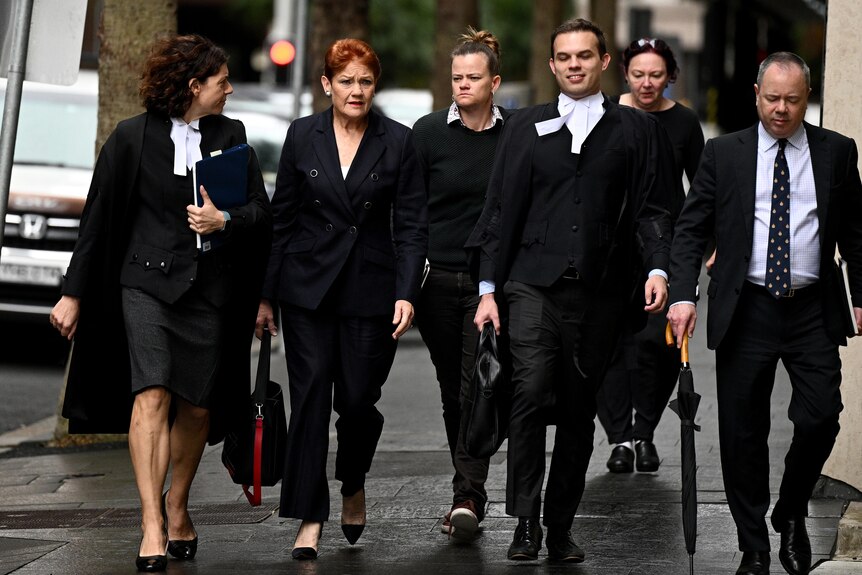 An older woman in all black walking with lawyers around her. 