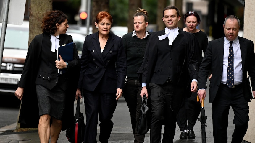 An older woman in all black walking with lawyers around her. 