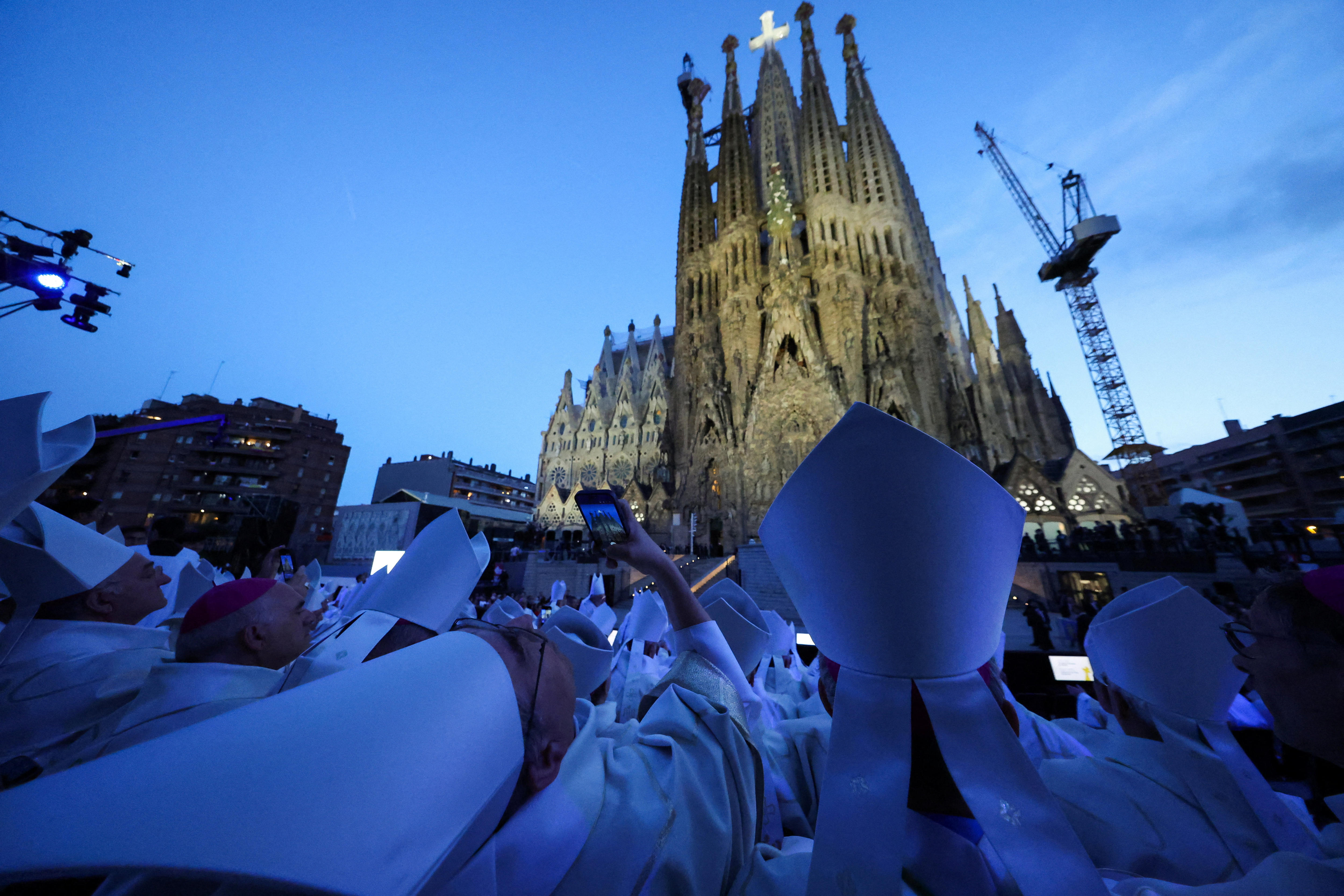 Catholic Church clergy members in white robes taking mobile phone photos at dusk of the exterior of the Sagrada Familia.