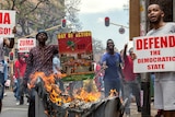 Protesters hold up signs reading President Zuma must go and another reading defend the democratic state. A fire burns in a bin