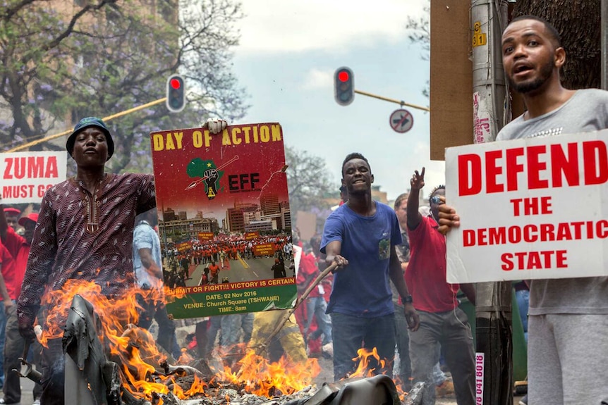 Protesters hold up signs reading President Zuma must go and another reading defend the democratic state. A fire burns in a bin