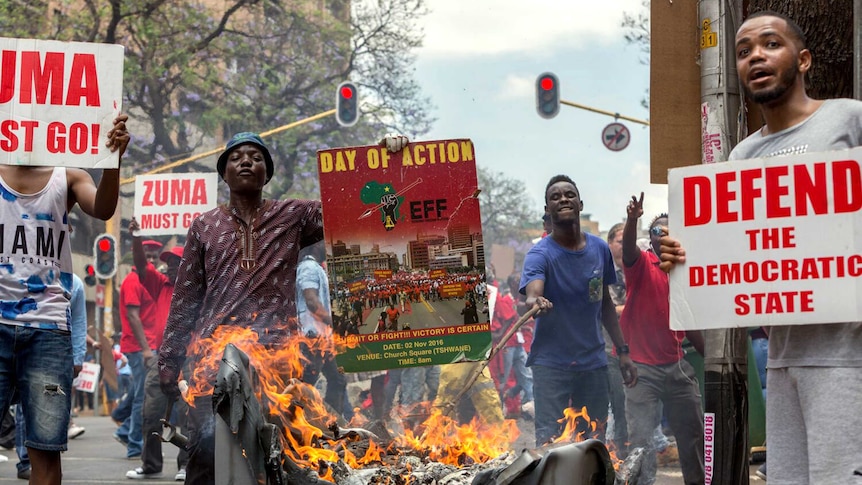 Protesters hold up signs reading President Zuma must go and another reading defend the democratic state. A fire burns in a bin