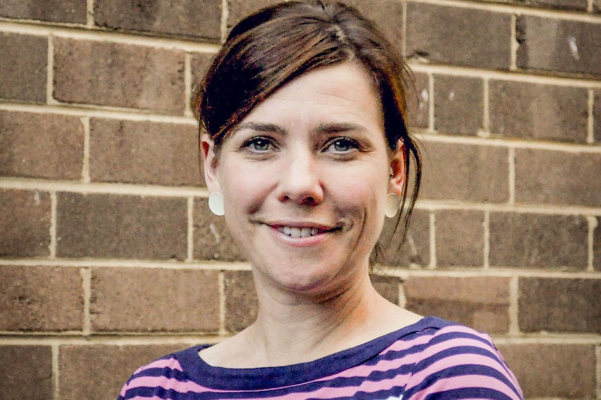 A young woman with dark hair smiles, standing near brick wall, wearing purple and white striped shirt.