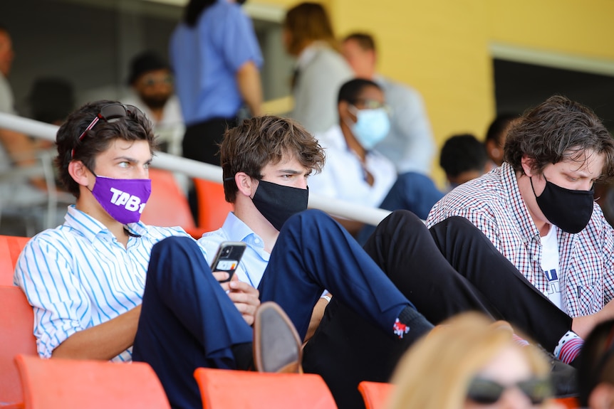Three young men sit together wearing masks at a horseracing event