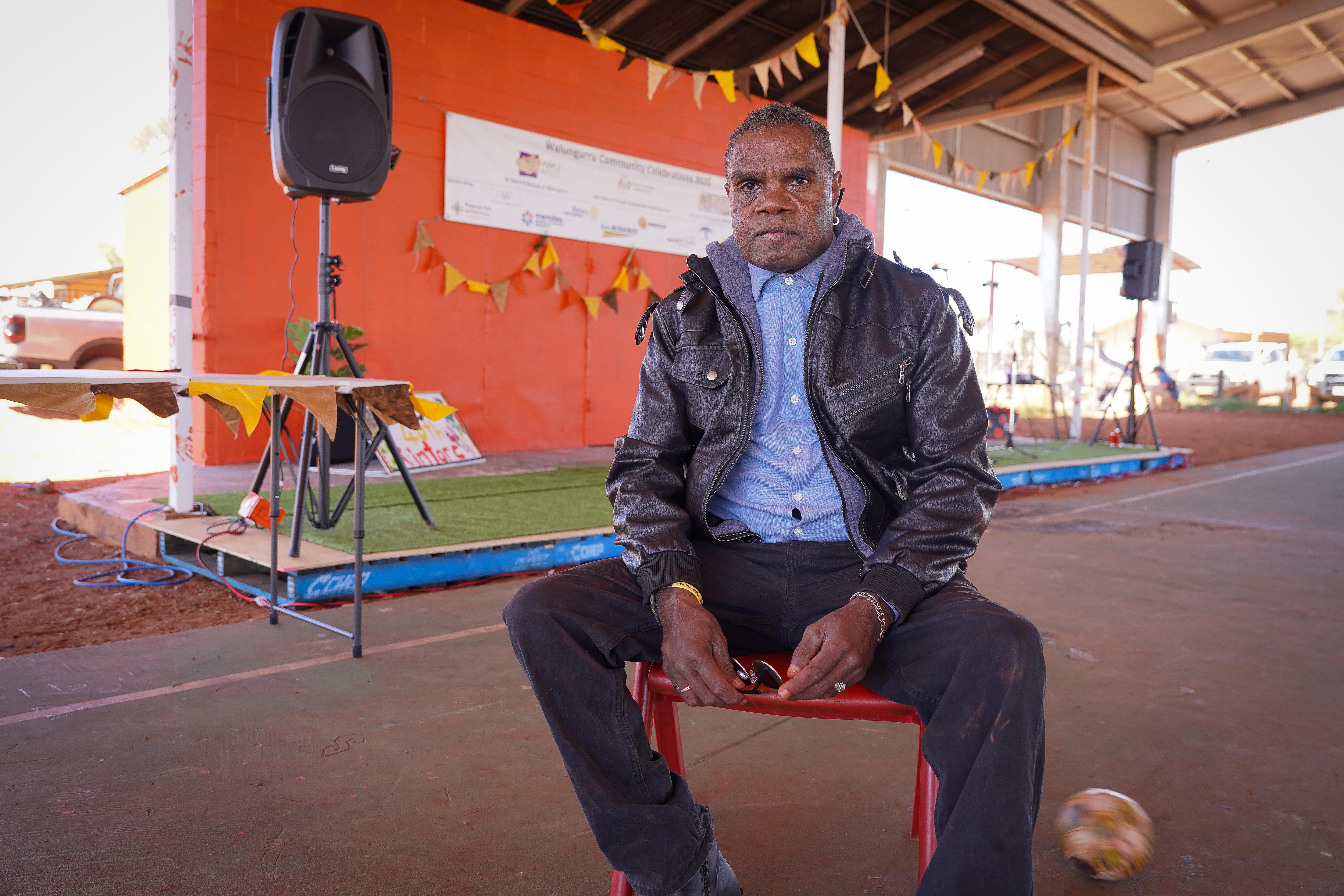 A middle-aged man in a leather jacket sits in freont of a low stage beneath a shade shelter.