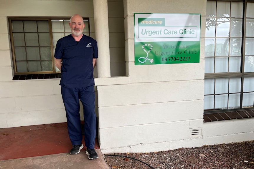 Un homme souriant et barbu en tenue de travail se tient devant une clinique médicale.