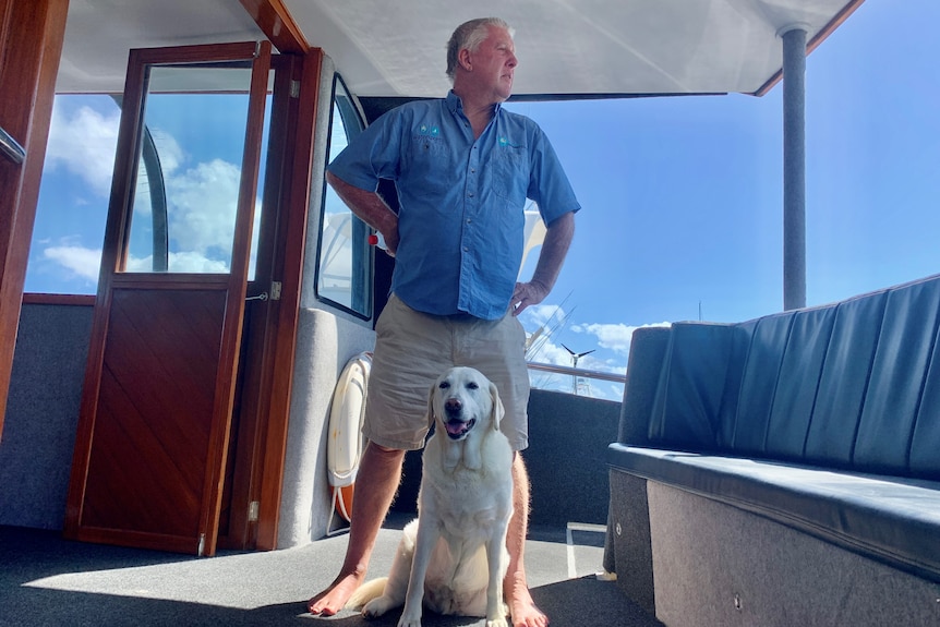 A shoeless man in beige shorts and blue shirt stands on a boat with a pale Labrador dog sitting in front of him