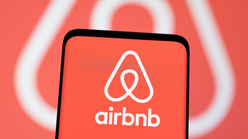A smartphone with an Airbnb logo on it in front of the same logo being projected on a screen.  