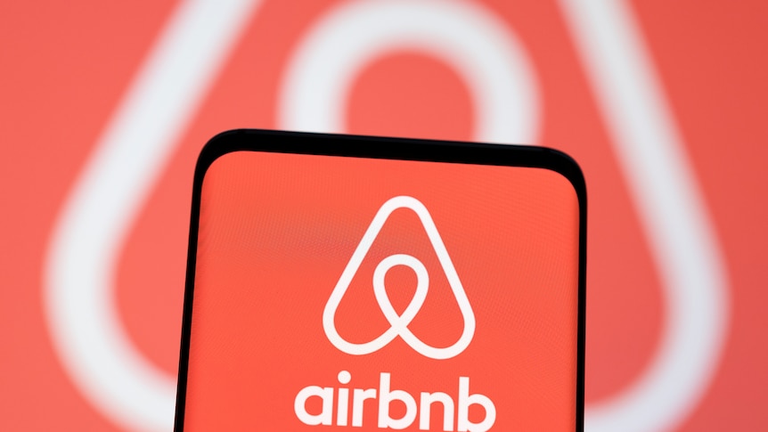 A smartphone with an Airbnb logo on it in front of the same logo being projected on a screen.  