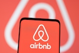 A smartphone with an Airbnb logo on it in front of the same logo being projected on a screen.  