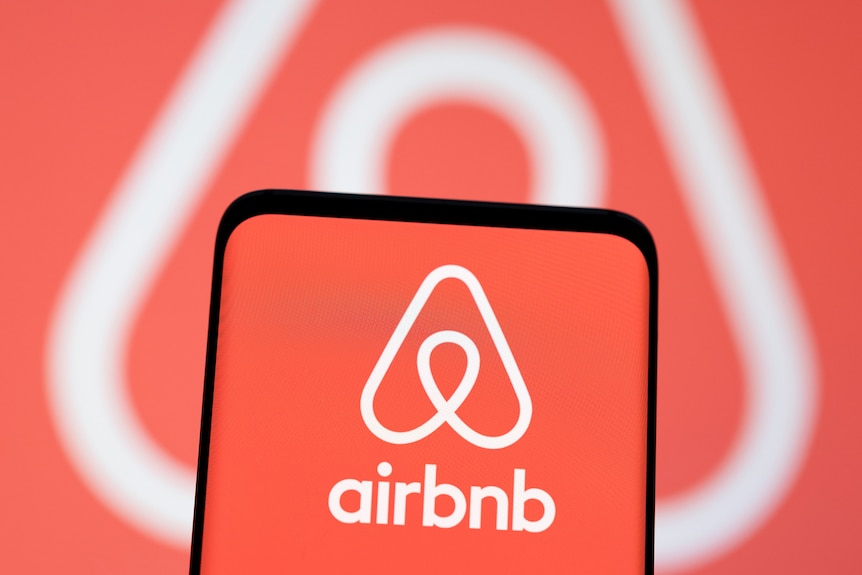 A smartphone with an Airbnb logo on it in front of the same logo being projected on a screen.  