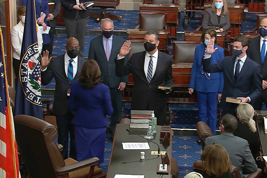 Three men in suits and face masks hold up their right hands while facing Kamala Harris.