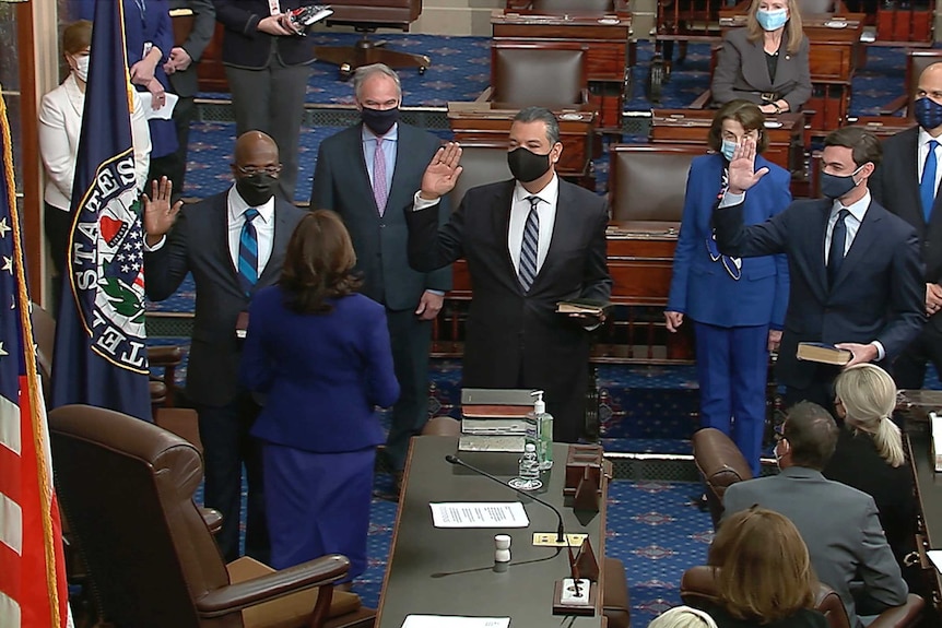 Three men in suits and face masks hold up their right hands while facing Kamala Harris.