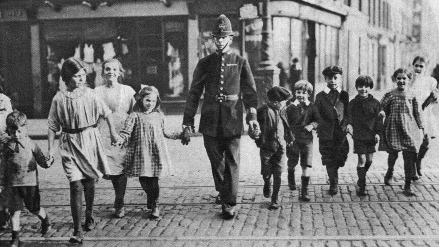 A black and white photo of a policeman helping children cross the road.