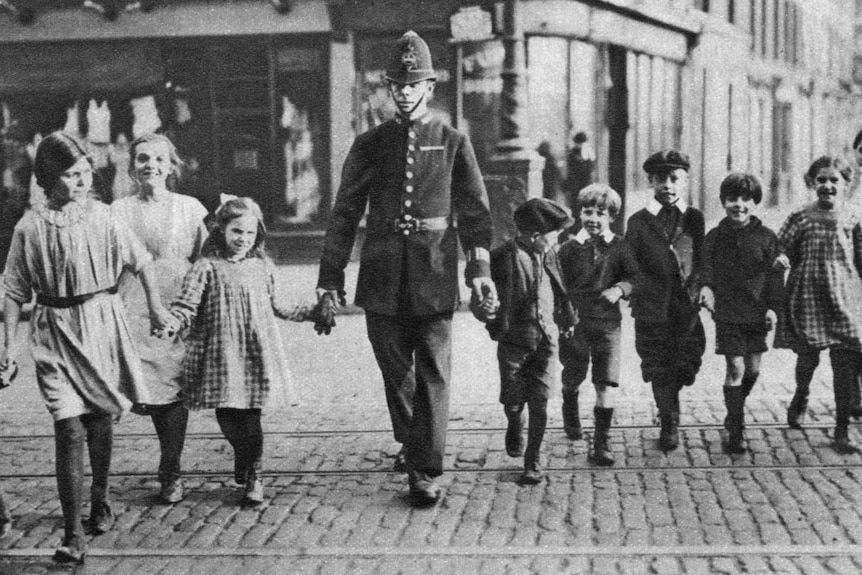 A black and white photo of a policeman helping children cross the road.