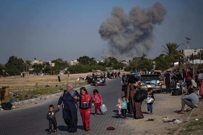 A mother with small children walks down a road with smoke from a bombardment seen in the village behind them.