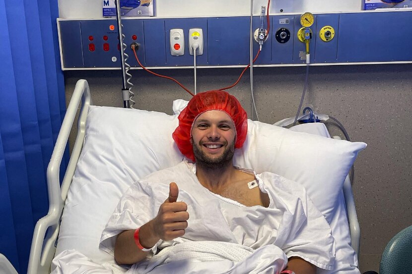 Benjamin Hampton in a hospital bed with blankets and a red surgical hairnet smiling and giving the thumbs up
