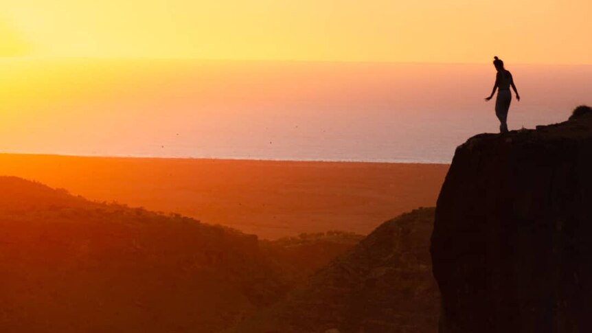 A person stands on the edge of a canyon at sunrise.