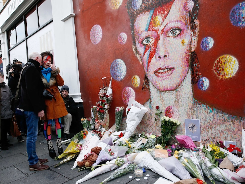A woman wearing Ziggy Stardust-style music cries next to a mural of David Bowie in Brixton