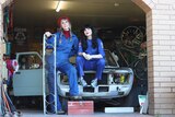 Two women in blue overalls sit and lean on car, one wears red beret and holds car grill.