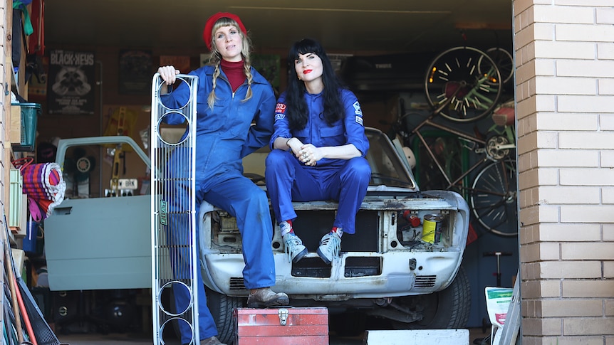 Two women in blue overalls sit and lean on car, one wears red beret and holds car grill.