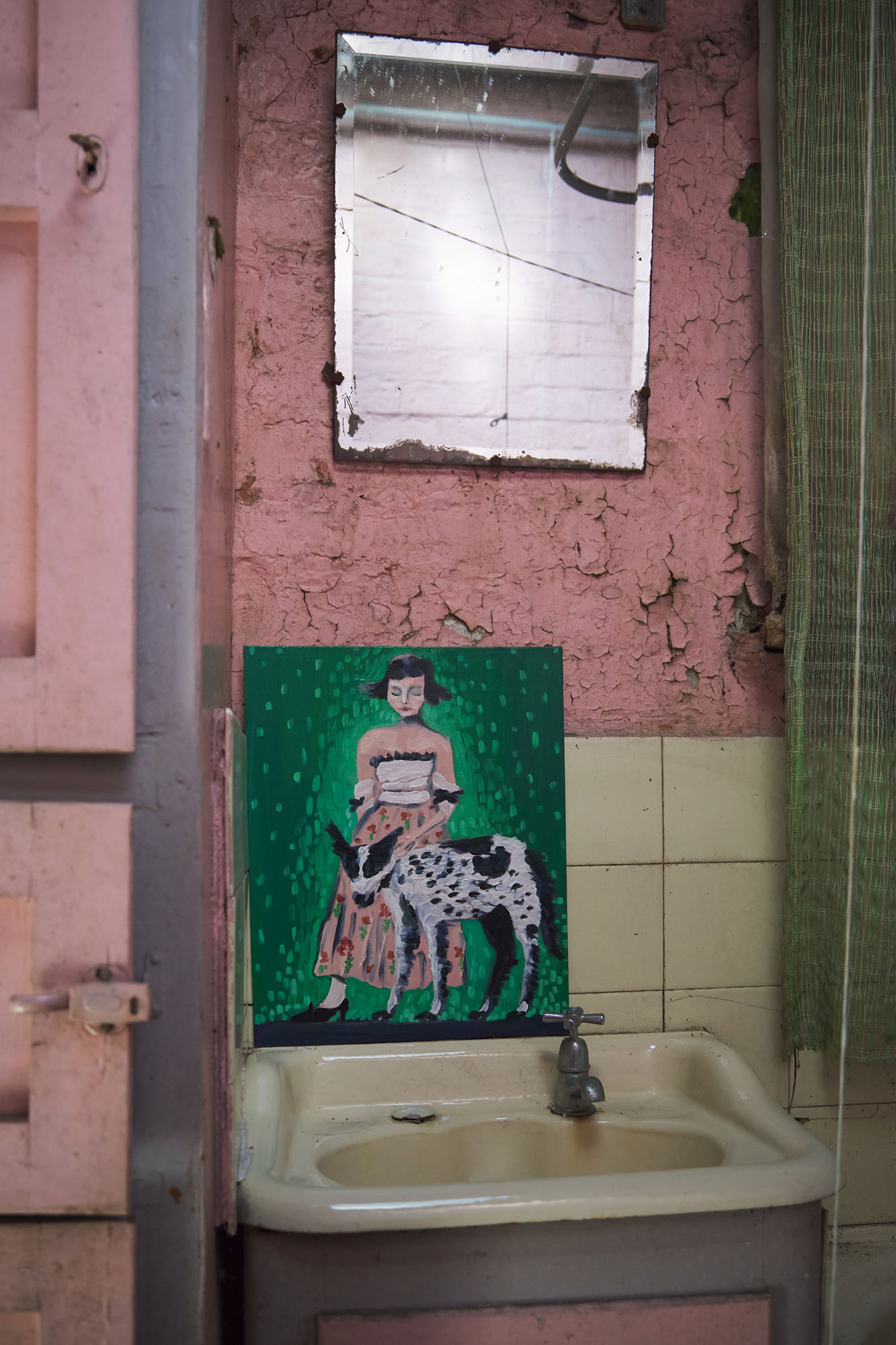 A decaying bathroom with peeling pink paint and a painting resting above the sink