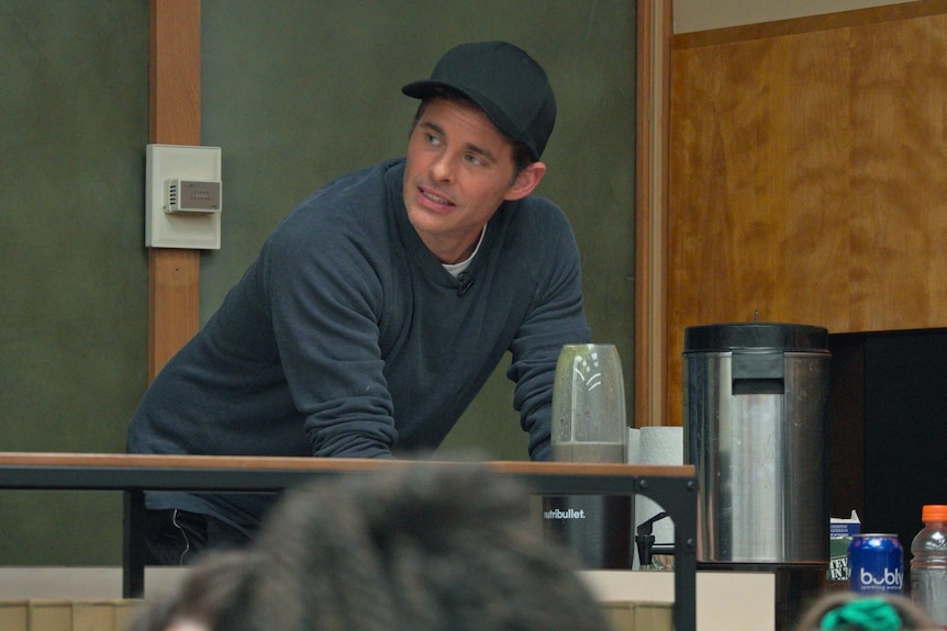 A man in his late 40s - the actor James Marsden - in a navy cap and long sleeved shirt, works a blender in a meeting room