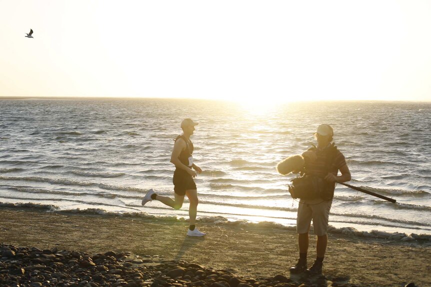 O'Brien running along beach past sound recordist holding boom microphone.