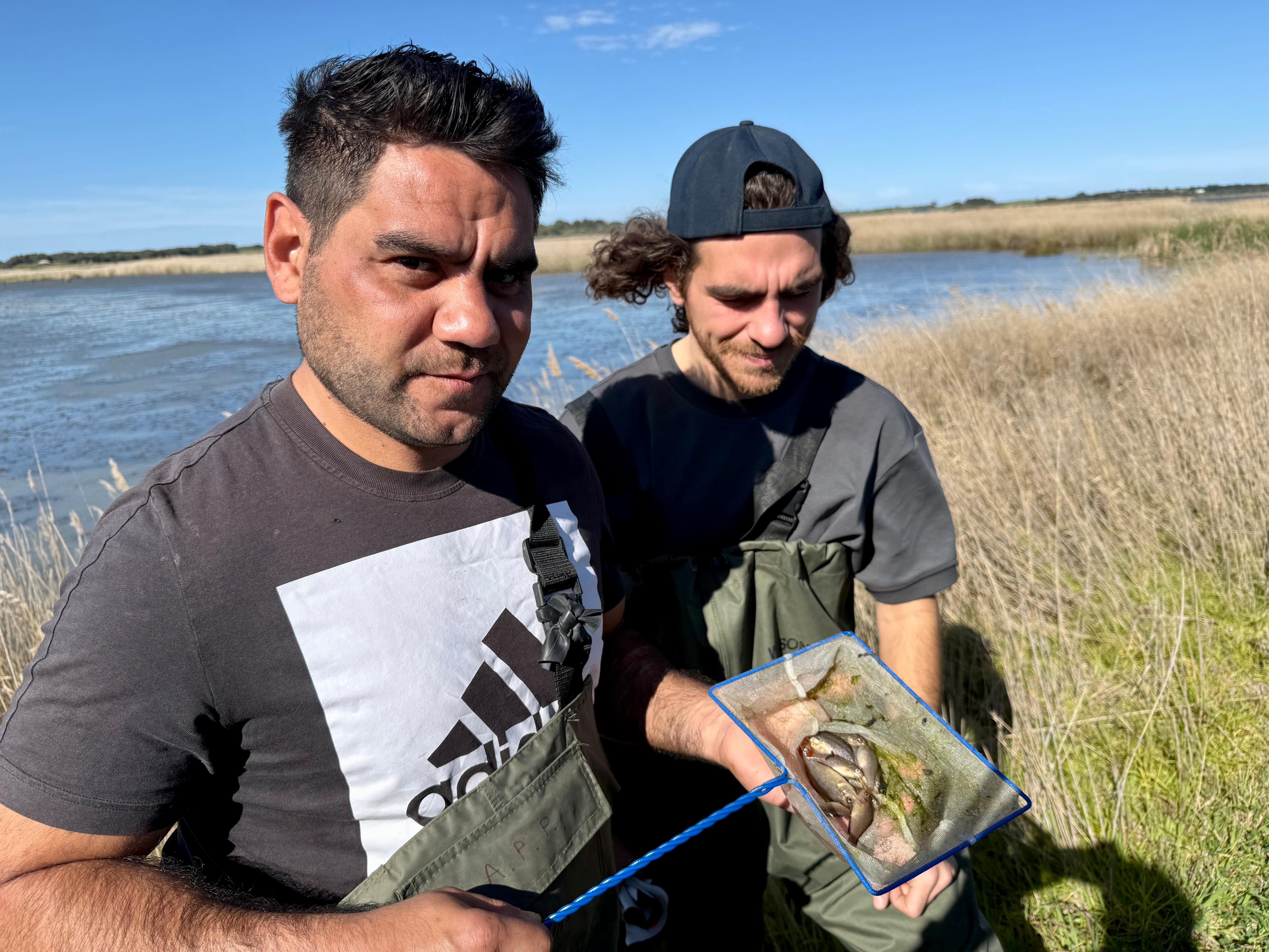 Two young men on a river bank with a small net full of fish.