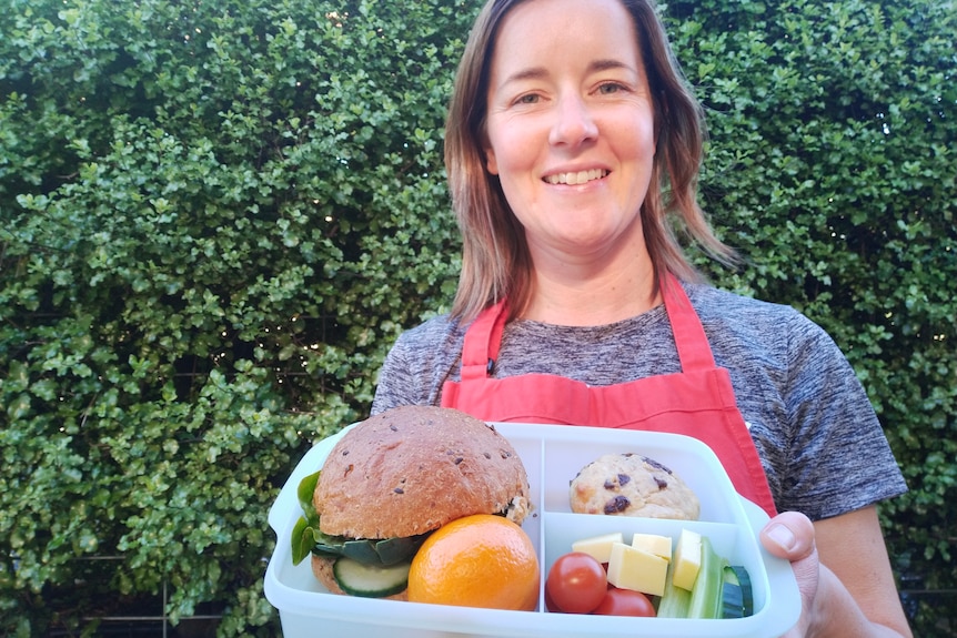 A smiling woman wearing an apron holds a lunchbox full of healthy food.