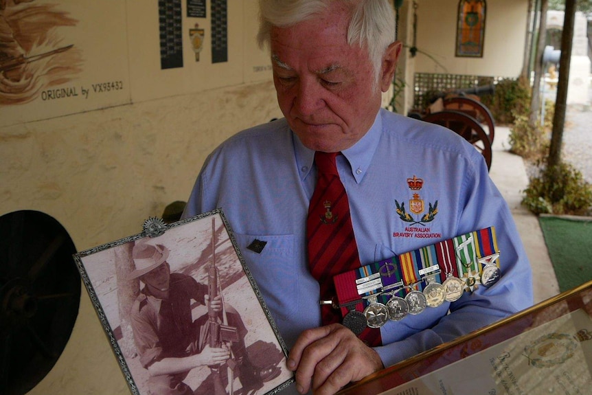 A man wearing military medals holds a photo of a soldier holding a gun
