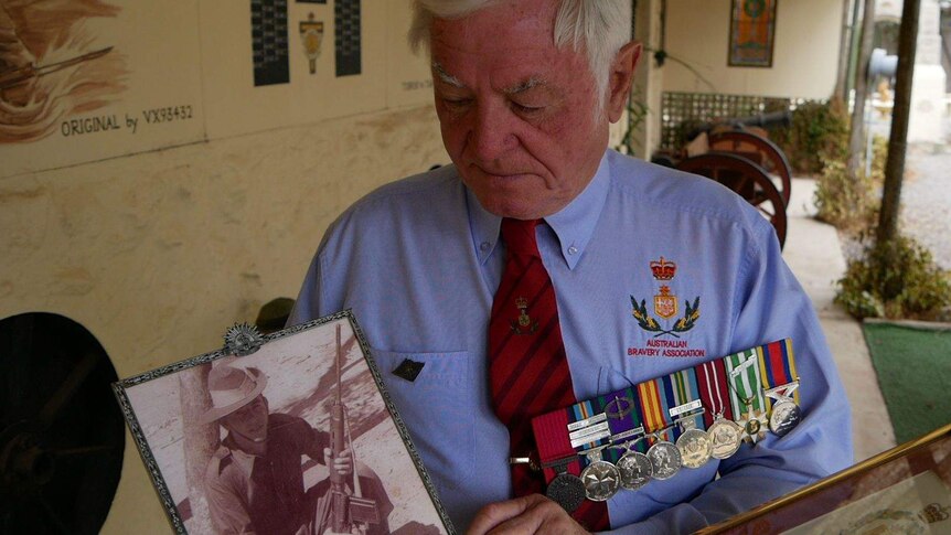 A man wearing military medals holds a photo of a soldier holding a gun