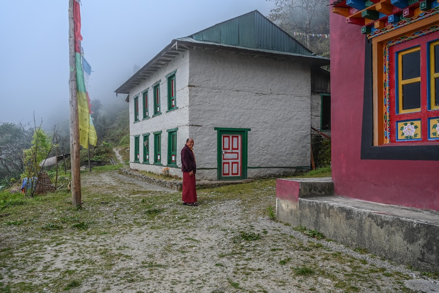 A man in robes stands outside a colourful building with mist in a valley behind