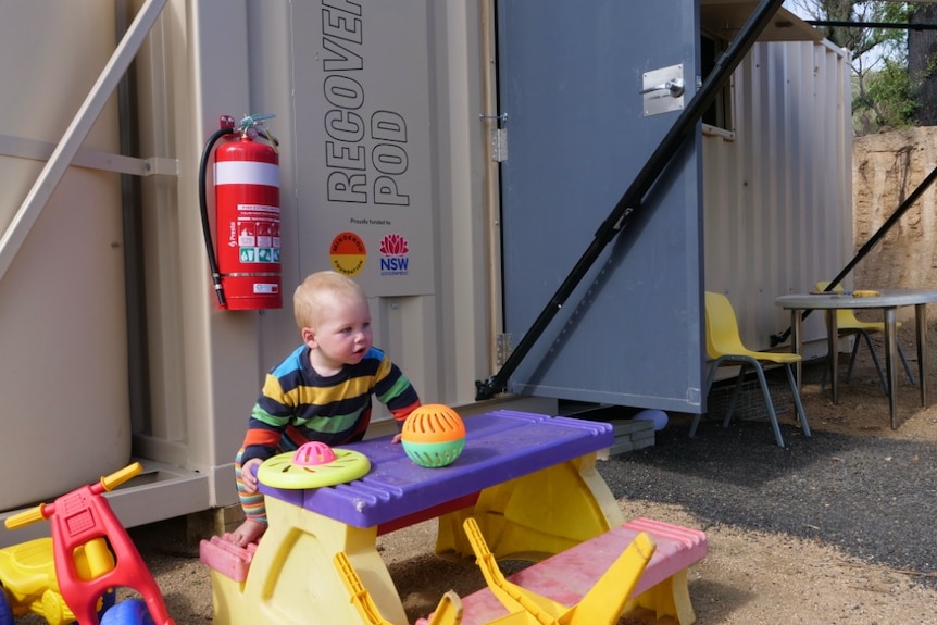 A little boy sits at a table outside a shipping container accommodation.