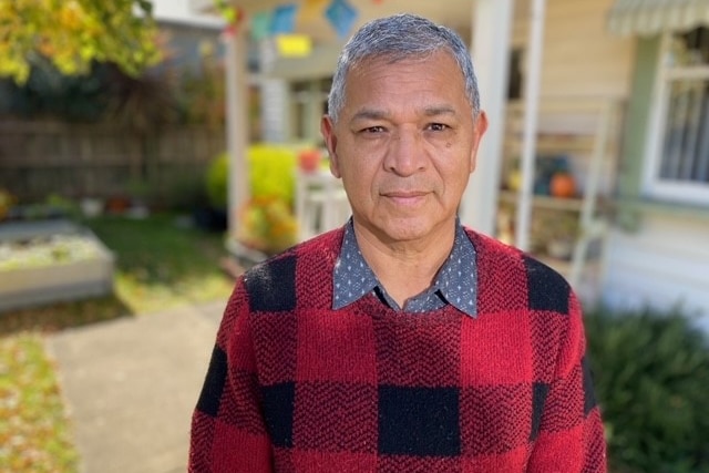An older man wearing a black and red checkered jumper stands outside a house on a sunny day.