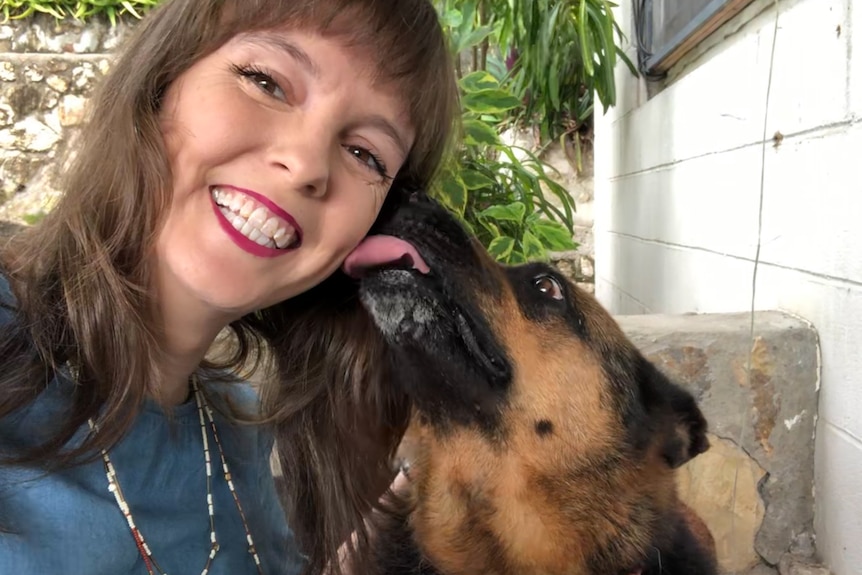 A great big German Shepherd licks a woman's face as she grins at the camera