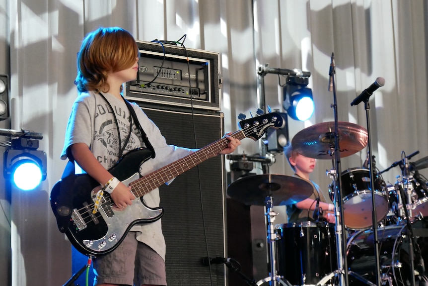 A young boy with long blonde hair holding a guitar on stage. Drums and lights behind.