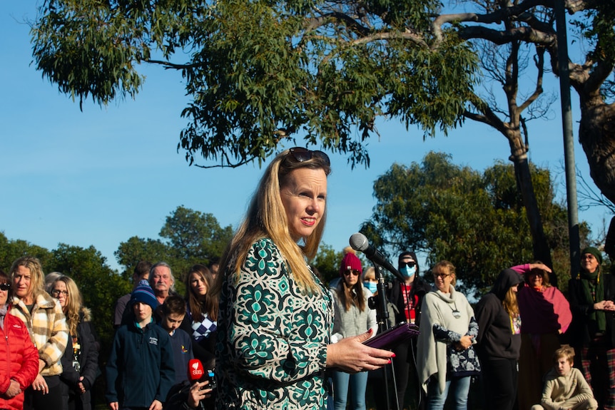 A woman with long blonde hair addresses a gathering outdoors.