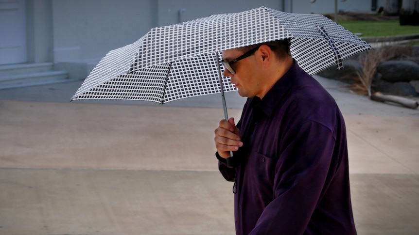 A in a dark jacket, wearing dark sunglasses walks while holding an umbrella over his head.